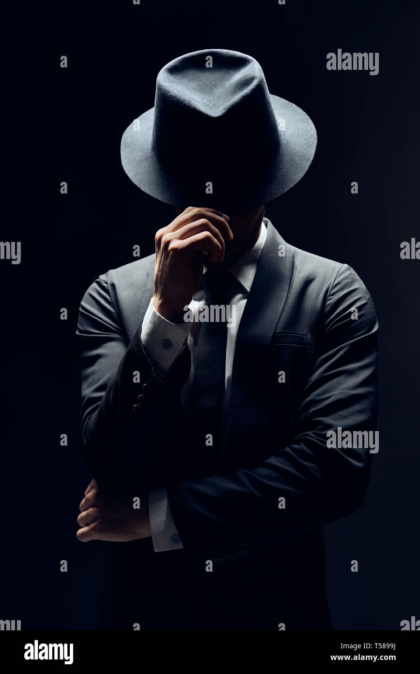 Mann in Anzug versteckt sich hinter seinen Hut isoliert auf dunklem Hintergrund. geheim und Incognito Konzept Stockfoto