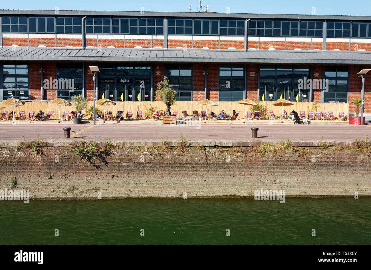 Streifen Sand, zwischen Gebäude und Straßen, Gras, Sonnenschirme, Liegestühle, Menschen entspannend, Seine Szene; Seawall, Wasser, humorvoll, einzigartige, Normand Stockfoto