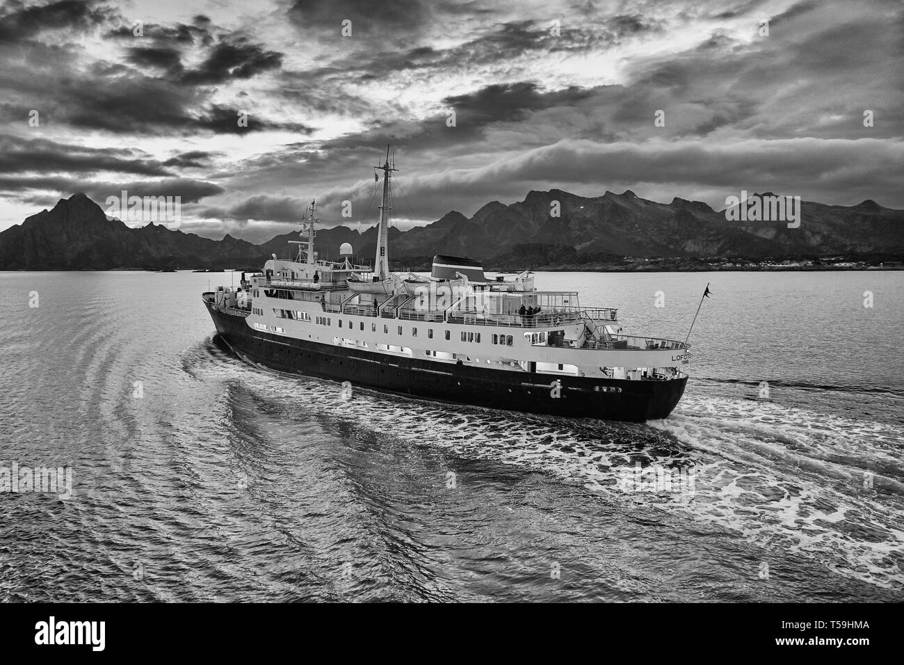 Ein Moody Black-White-Bild des klassischen Hurtigruten-Schiffs MS Lofoten, das im Vestfjord, den zerklüfteten Lofoten-Inseln dahinter, dampft. Norwegen. Stockfoto