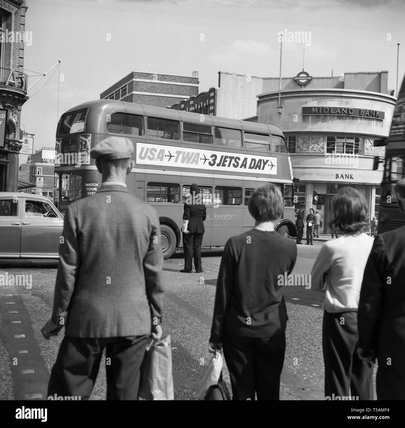 1960, historische, Menschen ein Polizist direkt beobachten den Verkehr als routemaster Bus versucht, biegen Sie links in eine Tube junction im Norden von London, um von einem Zweig des Midland Bank tun. Stockfoto