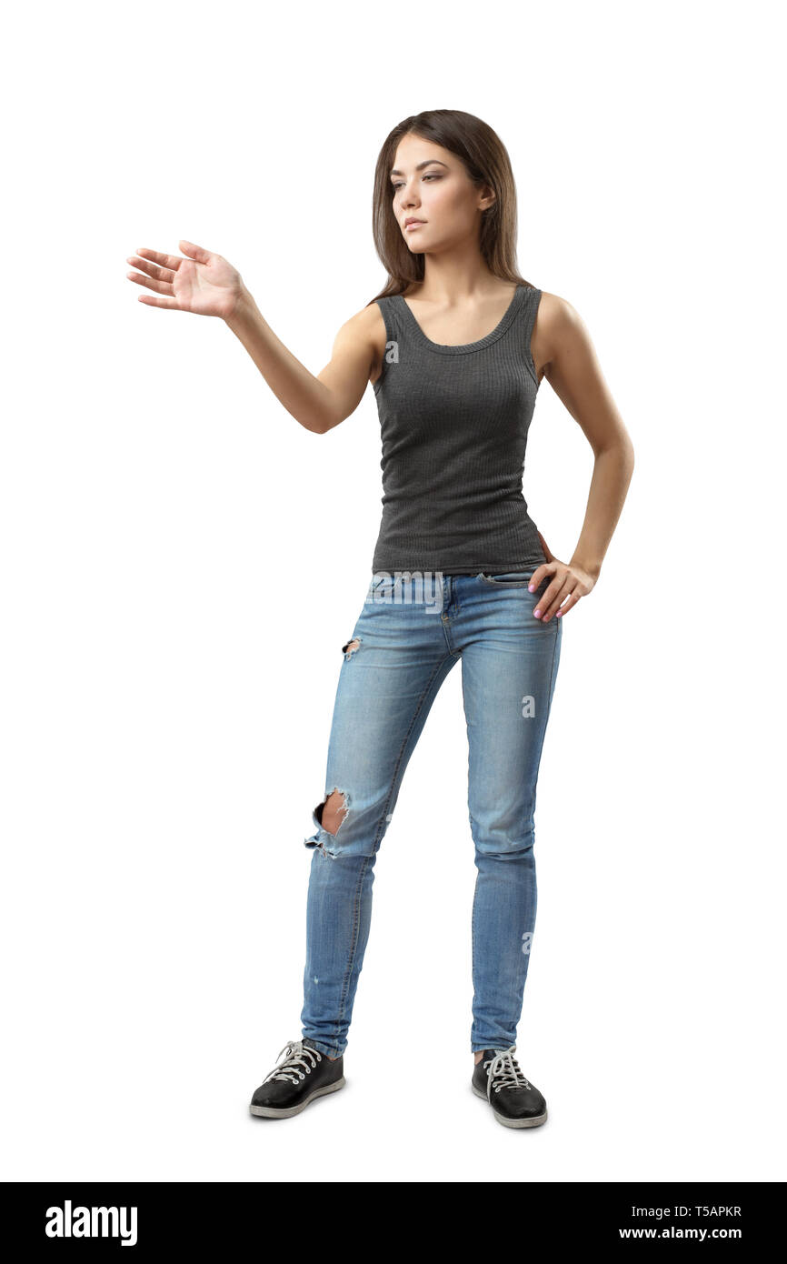 Junge Frau in Grau Top und Jeans stehend, eine Hand auf der Hüfte und andere in der Hand hielt, als wenn mit dem Finger auf dem Bildschirm unsichtbar auf weißem isoliert Stockfoto