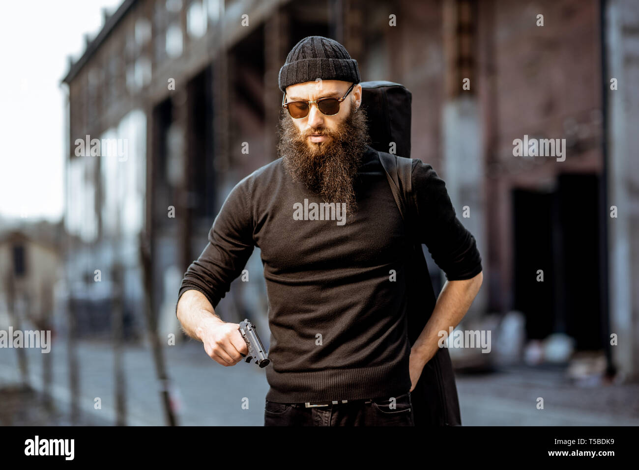 Ernsthafte bärtigen Mann in Schwarz feste Kleidung als Killer mit Pistole im Freien auf der industriellen städtischen Hintergrund gekleidet Stockfoto