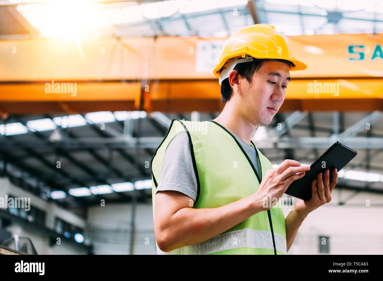 Jungen asiatischen Männlich schwere allgemeine Arbeitnehmer mit einem digitalen Tablet in Fertigung und Fabrikgelände mit Kran hinter Stockfoto