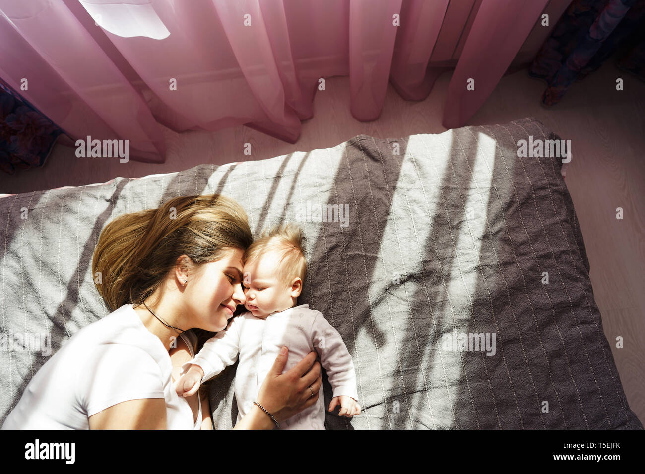 Mutter mit ihrem neugeborenen Sohn lag auf dem Bett in den Sonnenstrahlen aus dem Fenster durch die pinkfarbenen Vorhängen Stockfoto