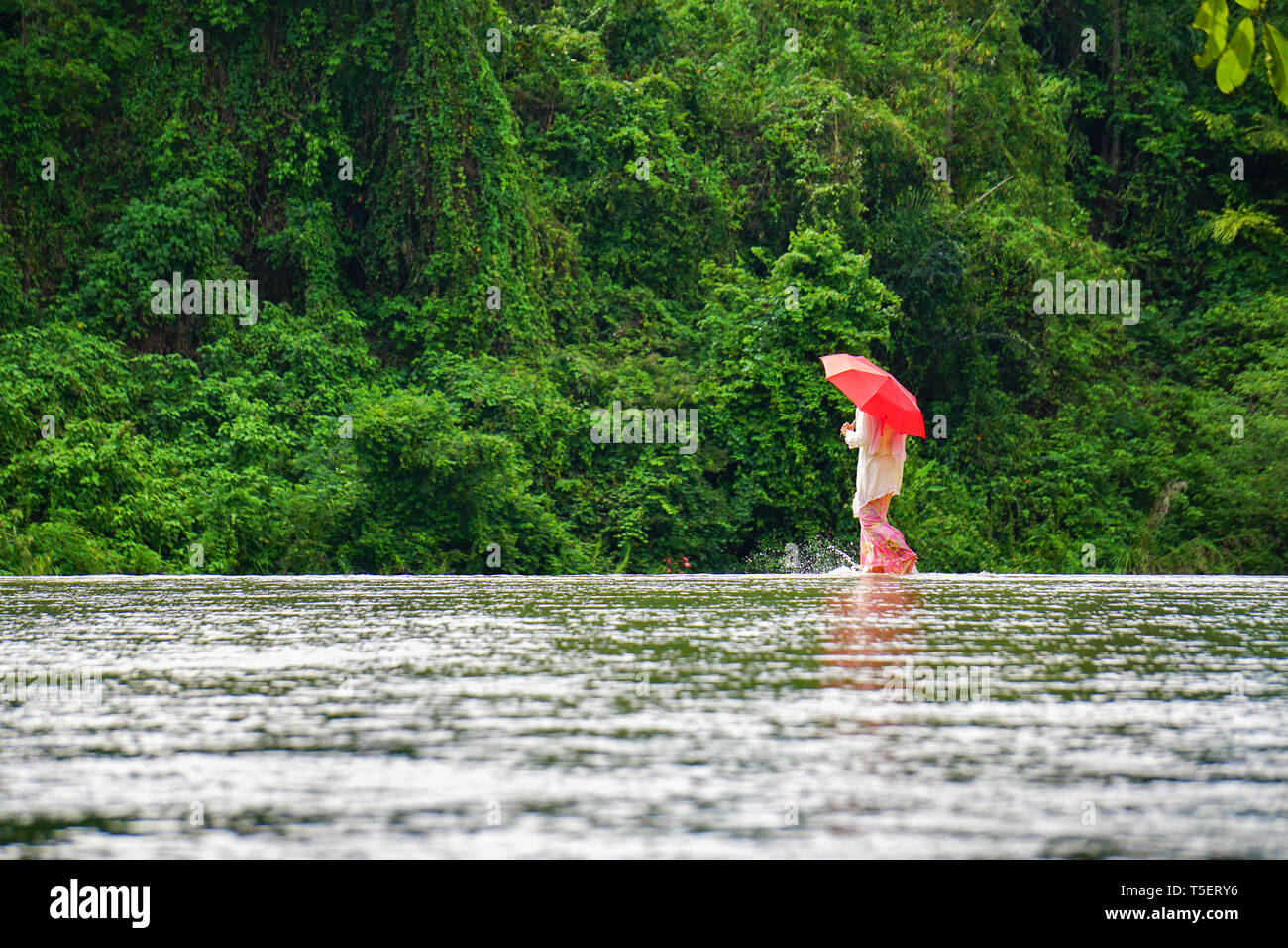 Dame in traditioneller Tracht Kebaya aufgerufen, mit roten Regenschirm zu Fuß auf River Crossing mit grünen Natur Hintergrund. Bild für Reisen geeignet Stockfoto