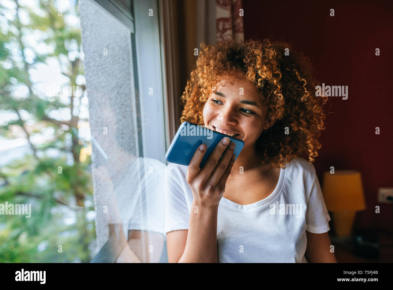 Junge Frau mit lockigem Haar Senden von Sprachnachrichten mit Handy neben dem Fenster Stockfoto