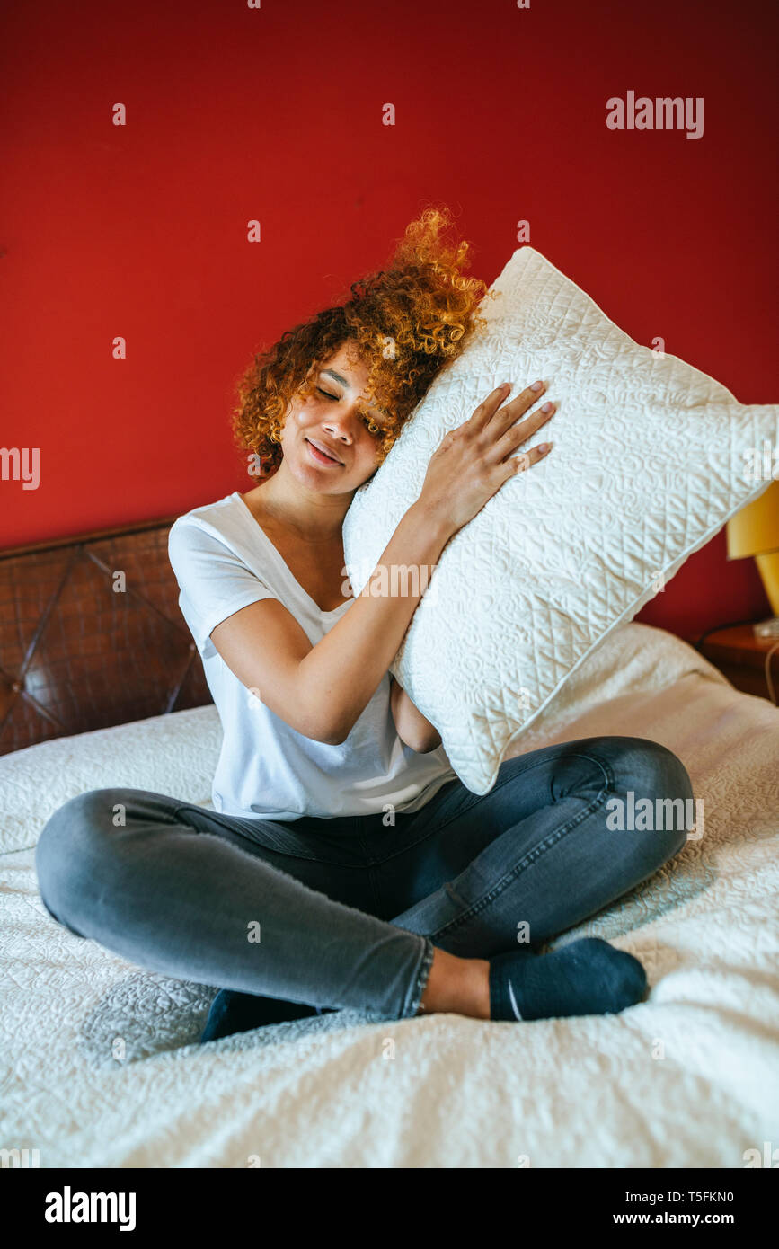 Junge Frau mit lockigem Haar saß auf dem Bett zu Hause holding Kissen Stockfoto
