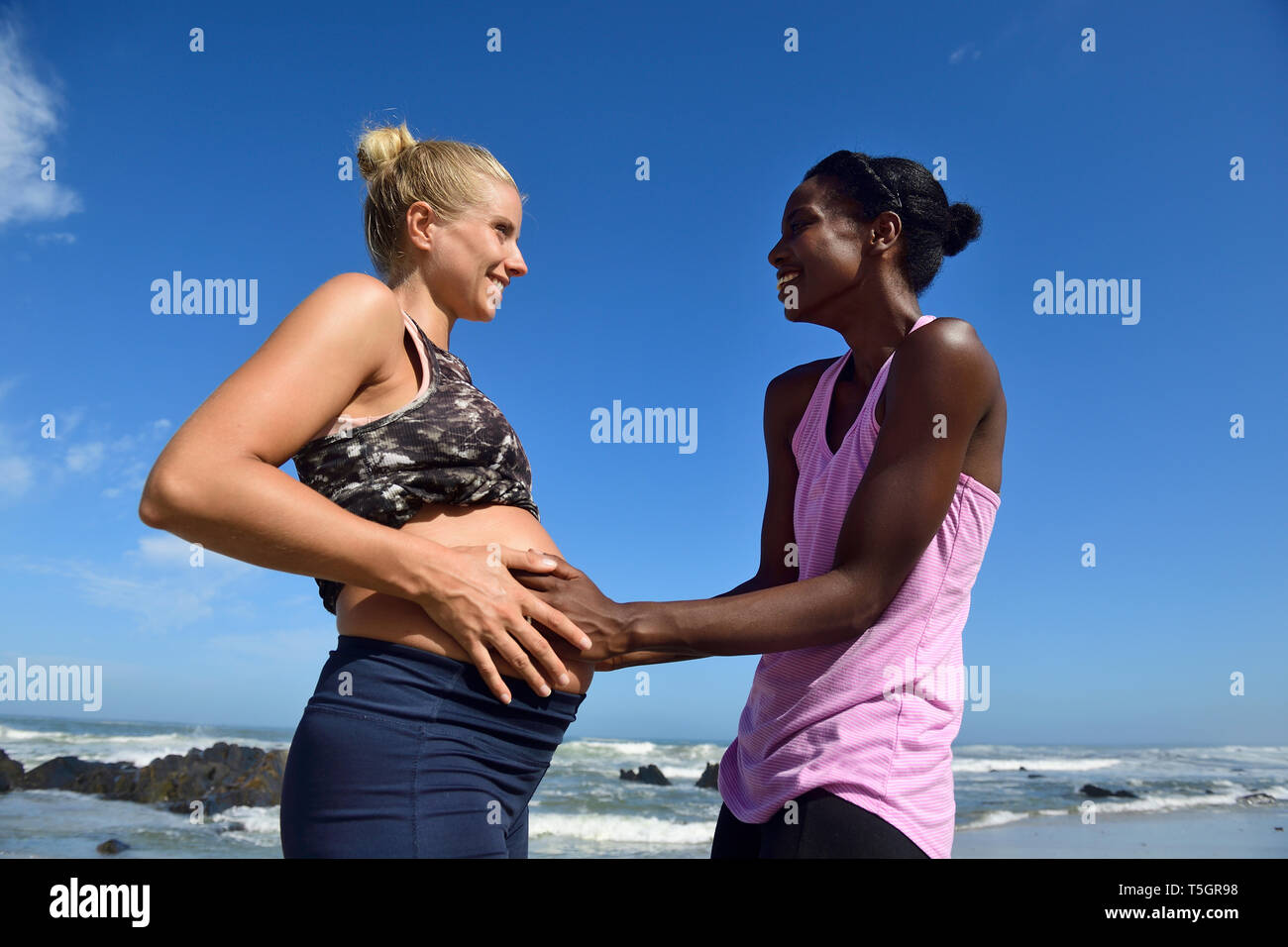 Glückliche Frau berühren Bauch der schwangeren Freundin am Strand Stockfoto