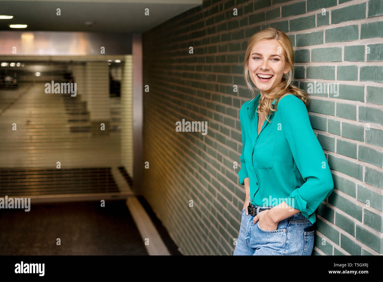 Portrait von eine fröhliche junge Frau, lehnte sich gegen die Wand mit den Händen in den Taschen Stockfoto