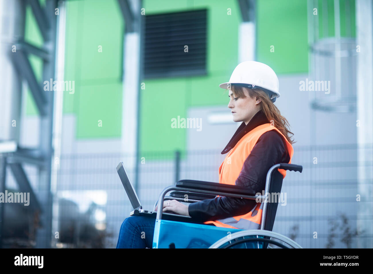 Junge Techniker mit Helm und Weste im Rollstuhl Arbeiten am Laptop im Freien Stockfoto