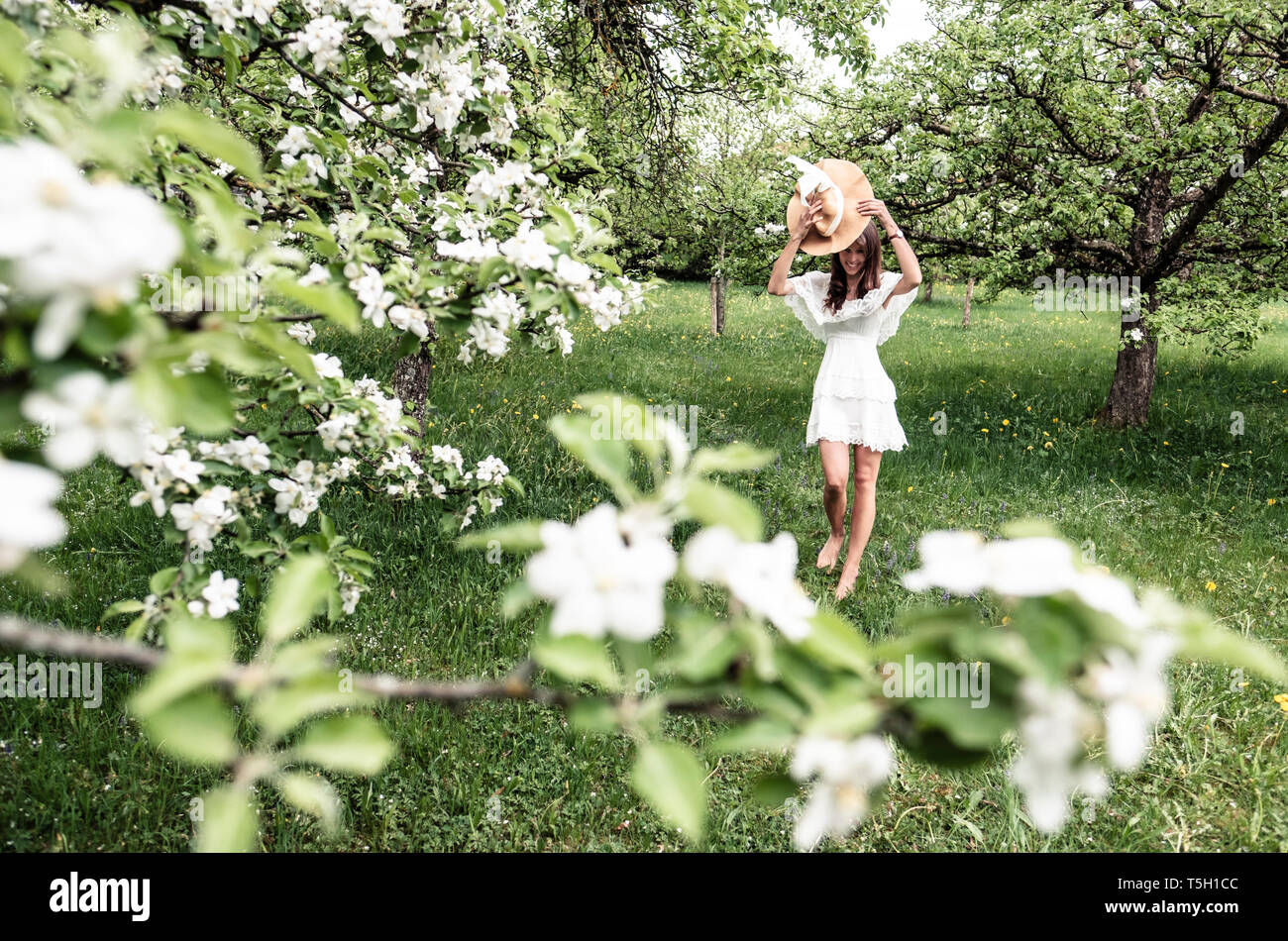 Junge Frau in weißem Kleid und Schlapphut barfuß im Garten mit blühenden Apfelbäume Stockfoto