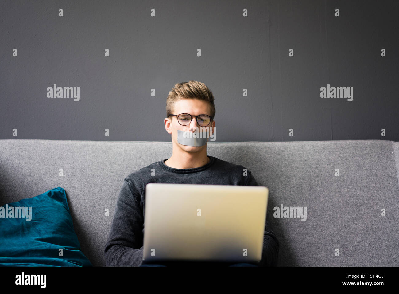 Junger Mann mit Getapten Mund sitzt mit Laptop auf der Couch Stockfoto