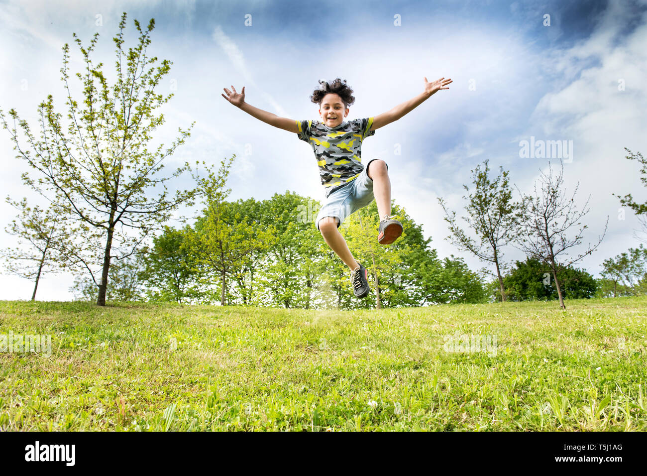 Happy energische junge Junge sprang hoch in die Luft, als er über das Gras in einem Garten oder Park mit ausgestreckten Armen in einem Low Angle View läuft Stockfoto