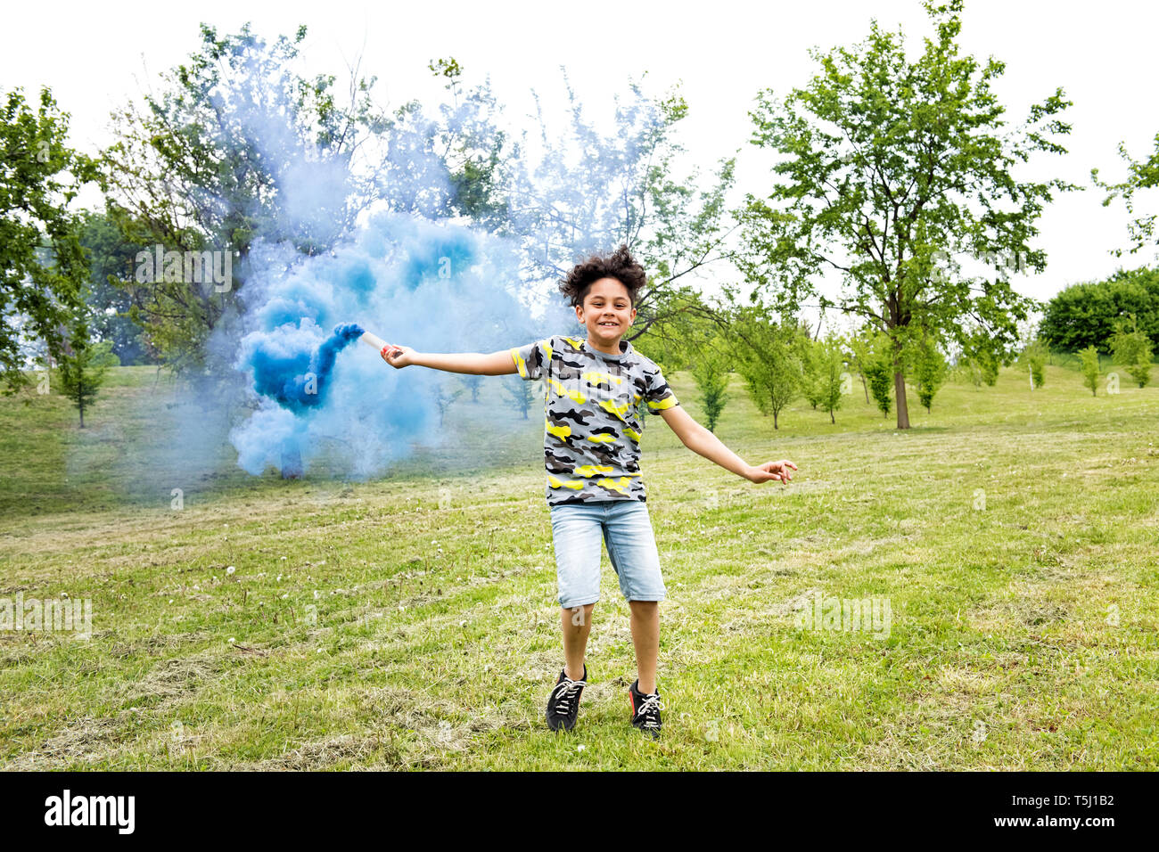 Junge schleppt ein blauer Rauch flare in einem Park über den rasen laufen mit einem strahlenden Lächeln der Freude und Genuss Stockfoto