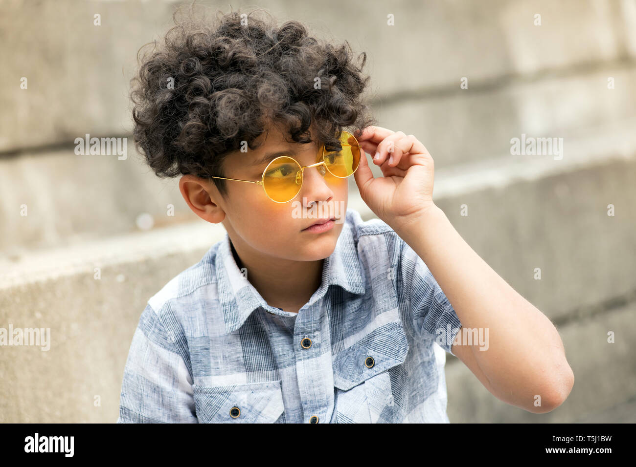 Junge curly vorangegangen Junge mit trendigen Gelber runder Schattierungen oder Sonnenbrille sitzen draußen auf der Seite betrachtet mit einem ernsten Ausdruck Stockfoto