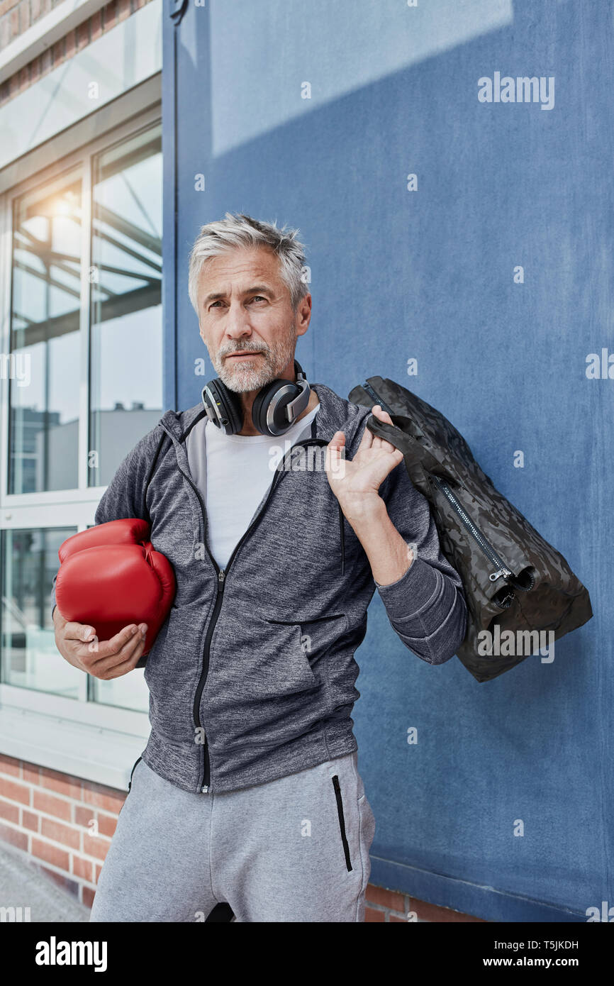Portrait von reifer Mann mit Kopfhörern, Sporttasche und roten Boxhandschuhe vor der Turnhalle stand Stockfoto