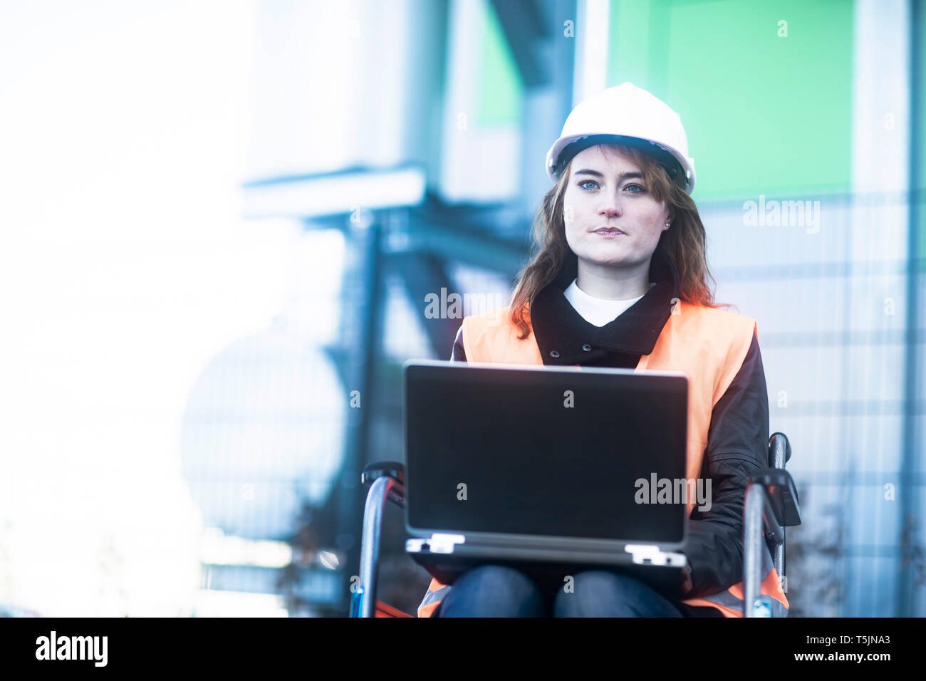 Porträt der jungen Techniker mit Helm und Weste im Rollstuhl Arbeiten am Laptop im Freien Stockfoto