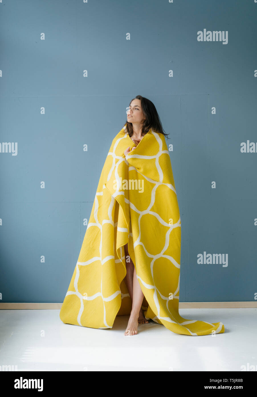 Frau stand vor der blauen Wand, in gelb Wolldecke eingewickelt Stockfoto