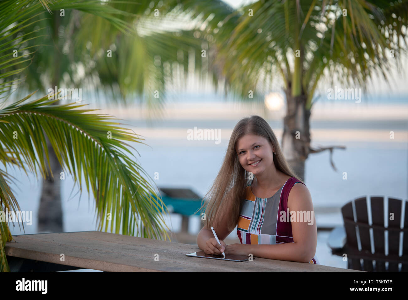 Junge Frau trägt mit Tablette am Strand morgens auf dem Hintergrund der Palmen in Florida Kleid Stockfoto