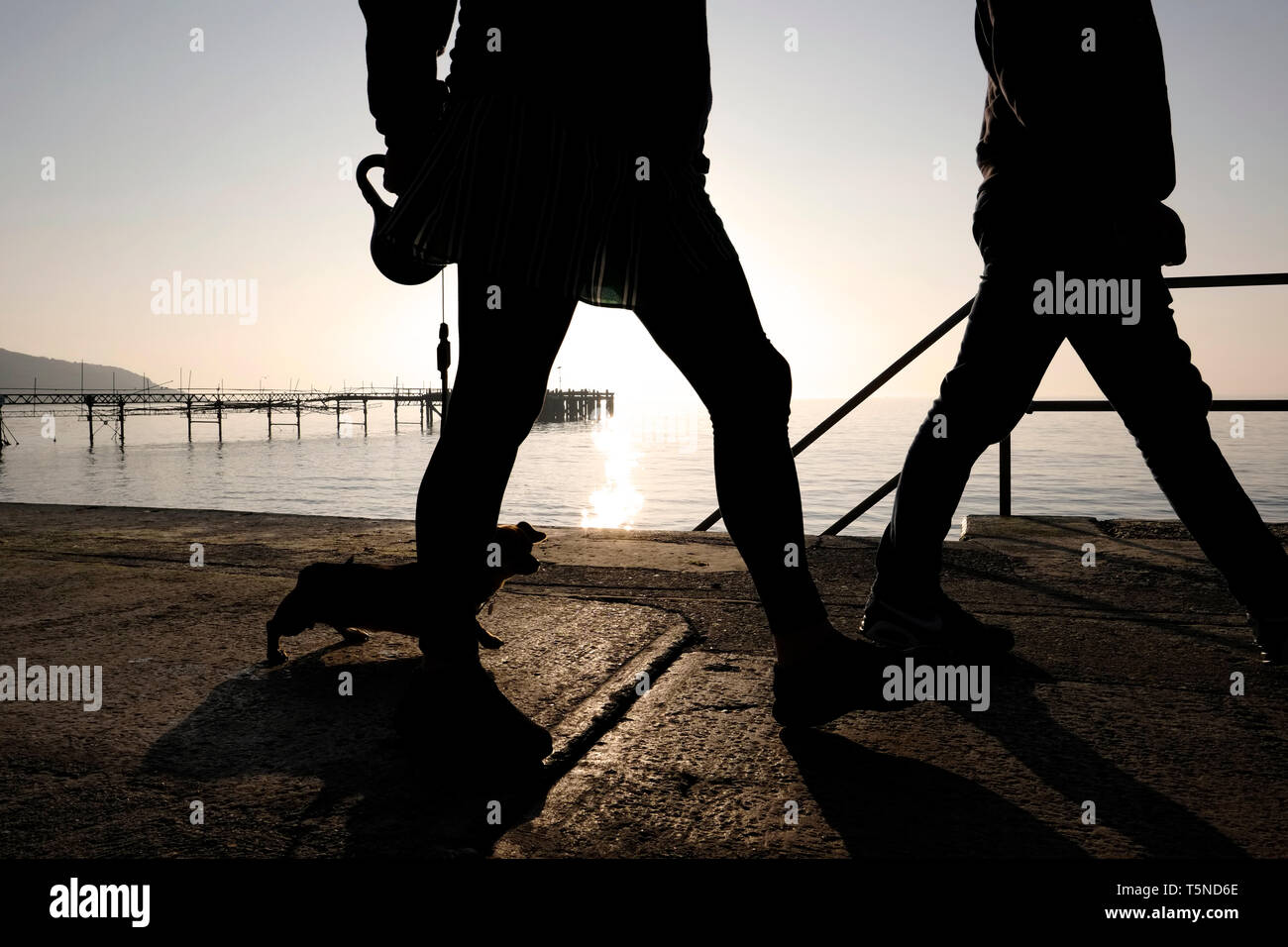 Spaziergang mit dem Hund entlang der Promenade Verkleidung Gehweg zwischen Totland und Colwell Buchten. Zwei Wanderer in Silhouette mit einer untergehenden Sonne über Totland Pier. Stockfoto