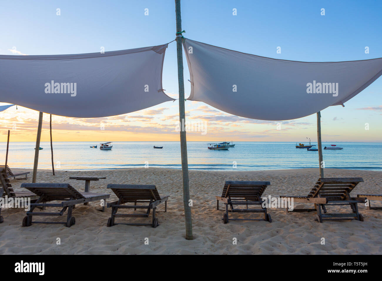Strand mit Stühlen aus Holz und Zelt mit Booten und den Sonnenuntergang im Hintergrund. Konzept der Ferien, Ruhe und Entspannung. Ponta do Corumbau, Bahia, Brasilien Stockfoto