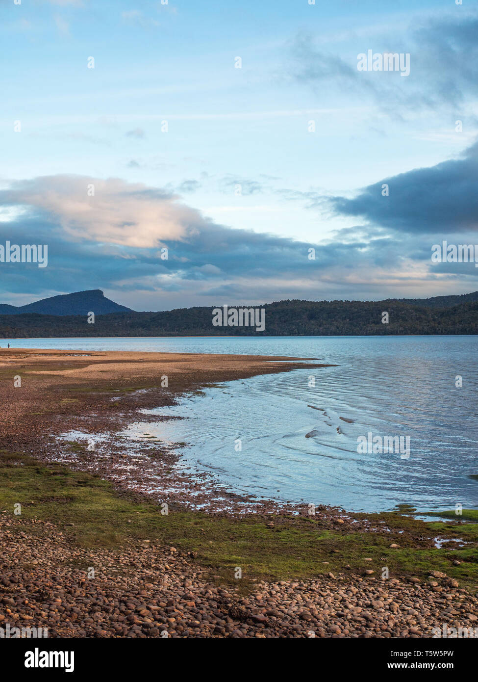 Kurve eines Pebble Beach, ein entfernter in ruhigem Wasser, von hohen Hügeln, unter Wolken in einen klaren Himmel, Lake Hauroko, Fiordland National Park, South Stockfoto