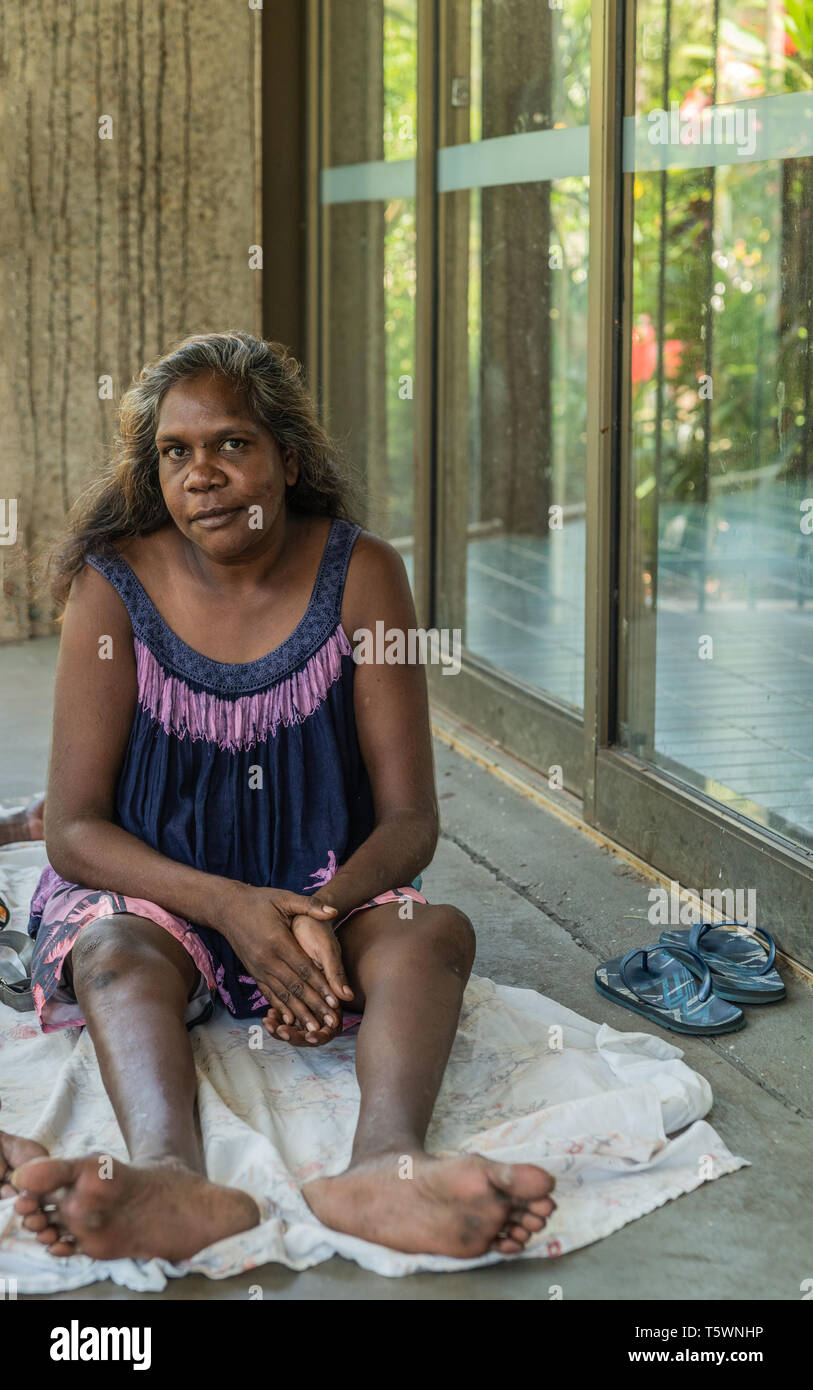 Darwin Australien - Februar 22, 2019: Nahaufnahme von jungen aboriginal Frau hinter im Schatten der Gebäude. Blau und Rosa Kleid, barfuß. Stockfoto