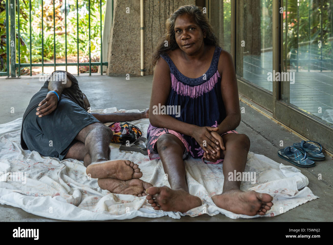 Darwin Australien - Februar 22, 2019: Nahaufnahme von jungen aboriginal Frau hinter im Schatten der Gebäude. Blau und Rosa Kleid, barfuß. Sle Stockfoto