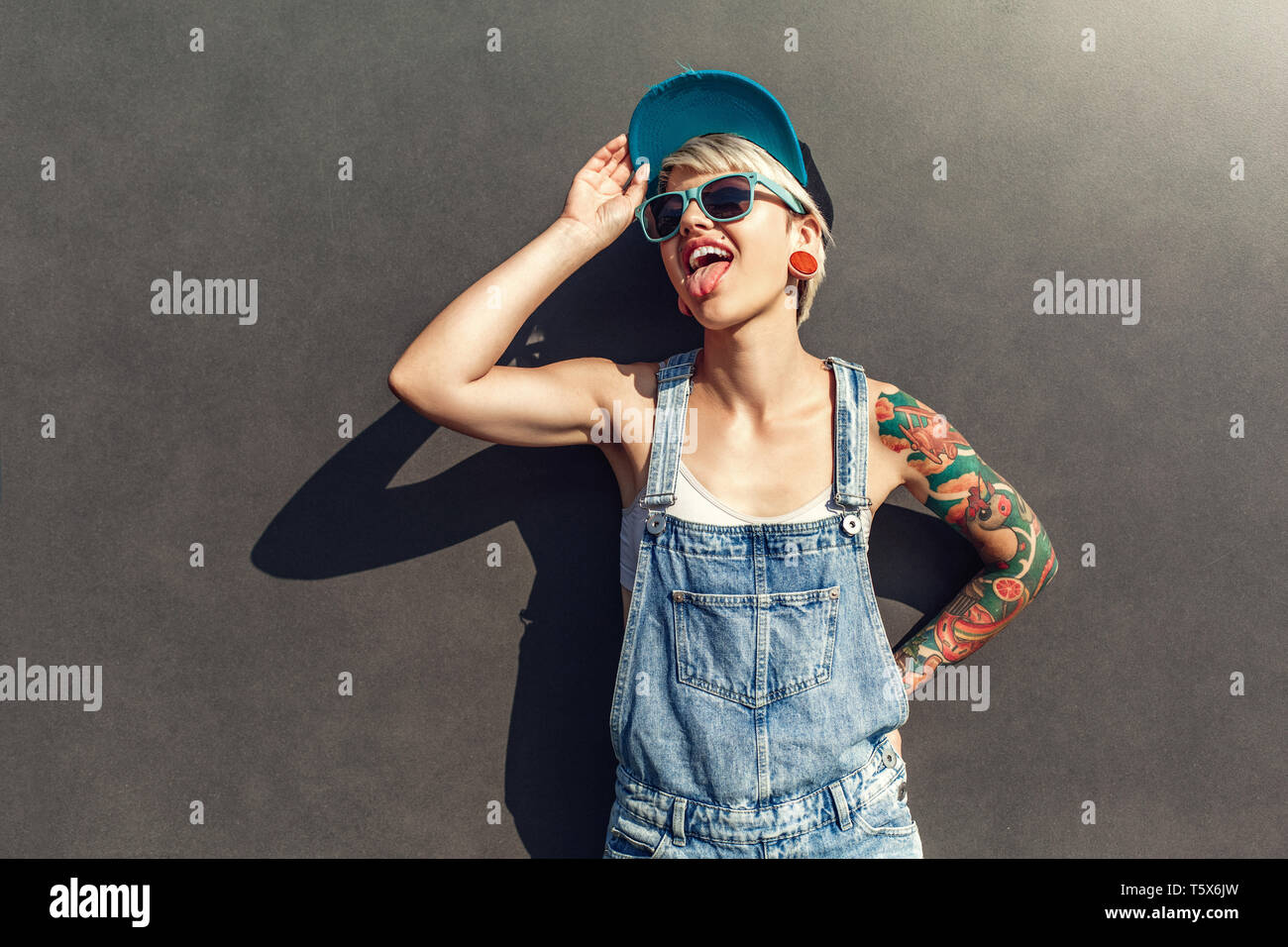 Junge alternative Mädchen mit Hut und Sonnenbrille auf der grauen Wand auf der Straße der Stadt isoliert stehende beiseite Suchen Übersicht Zunge verspielten Stockfoto