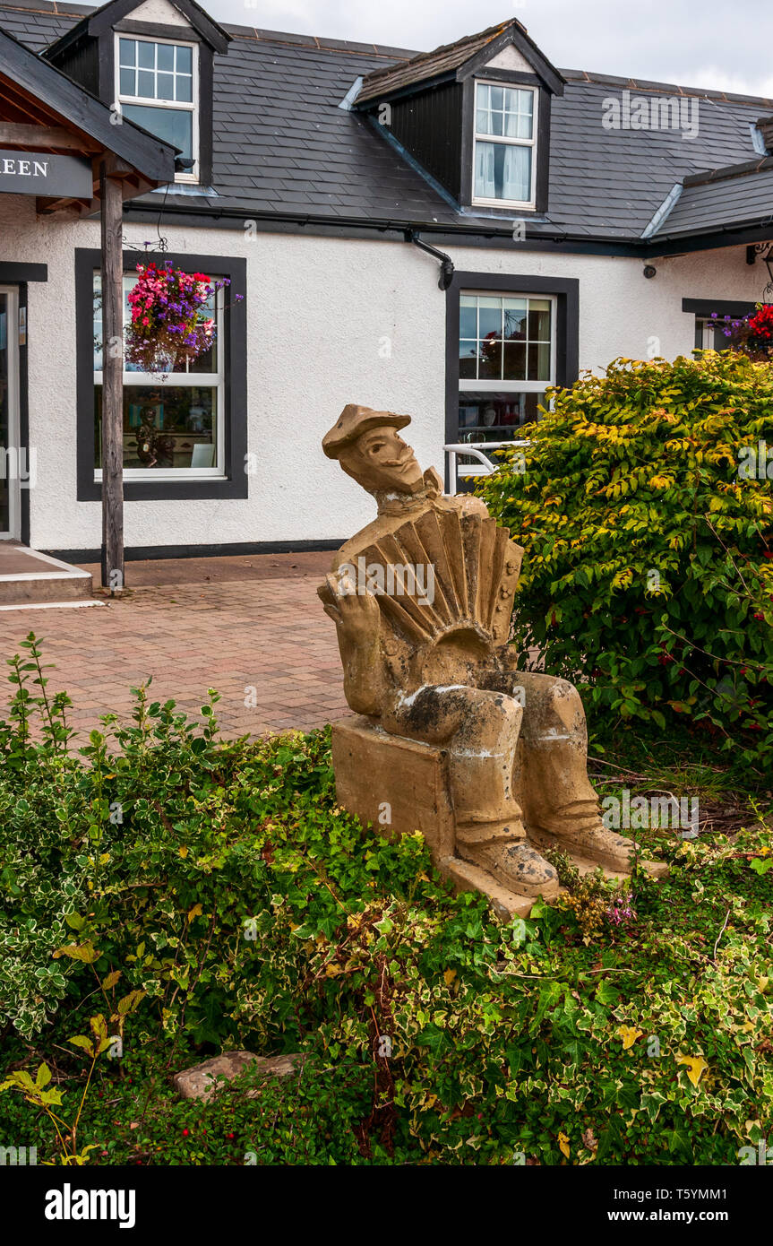 Eine braune Skulptur einer sitzenden Mann, lächelte, als er das Klavier Akkordeon spielt, zwischen zwei Büschen am Rand der Terrasse sitzen vor einem weißen Shop Stockfoto