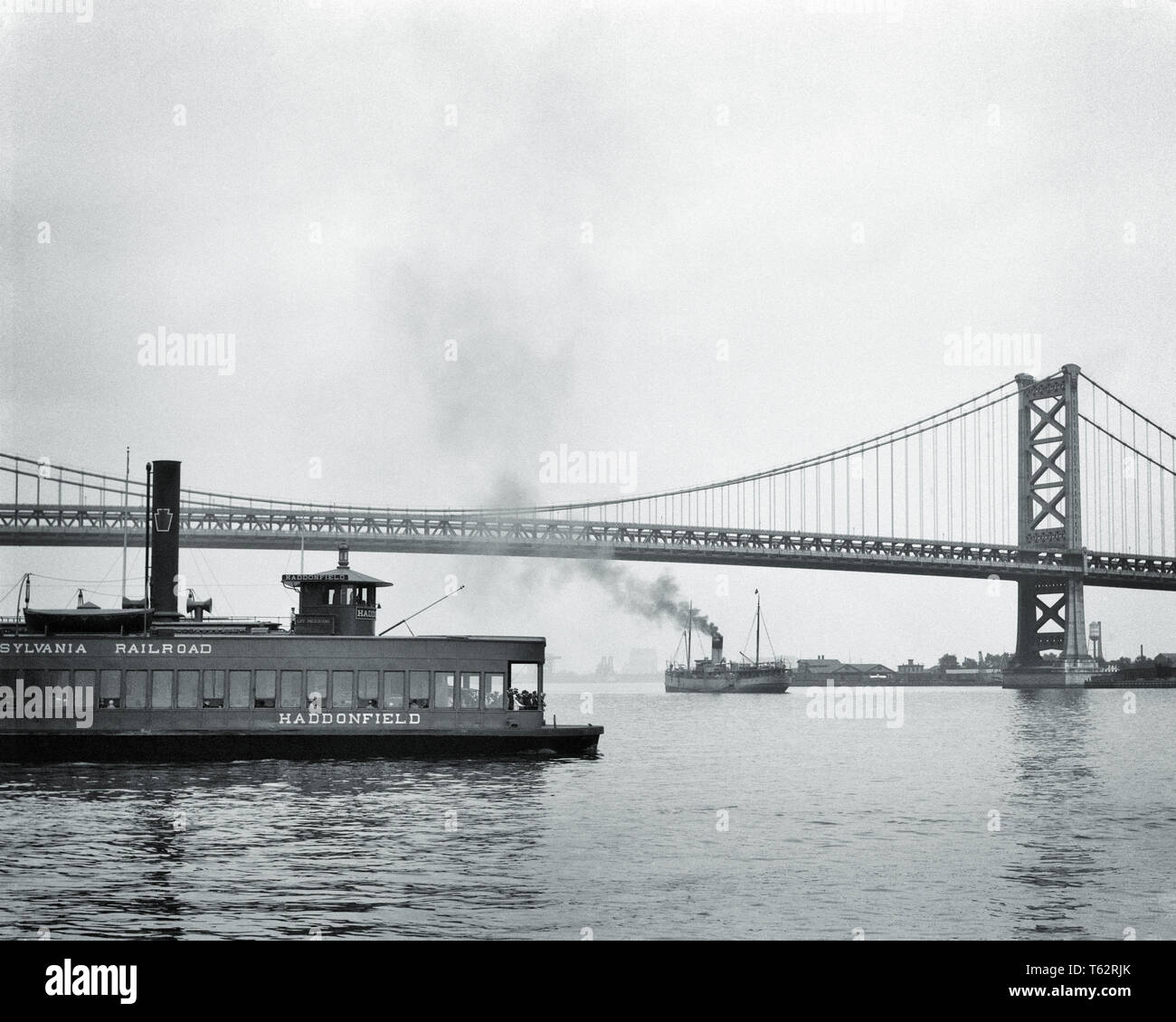 1920 Pennsylvania Railroad Fähre überqueren Delaware River new jersey Benjamin Franklin Bridge im Hintergrund haddonfield-b 3498 HAR 001 HARS FÄHREN NEW JERSEY BENJAMIN FRANKLIN BRIDGE ZUSAMMENARBEIT DELAWARE RIVER HADDONFIELD SPAN TRANSIT SCHWARZEN UND WEISSEN BRÜCKEN Stadt der brüderlichen Liebe HAR 001 MELDEN SIE ALTMODISCHEN DAMPFSCHIFF SUSPENSION BRIDGE Stockfoto