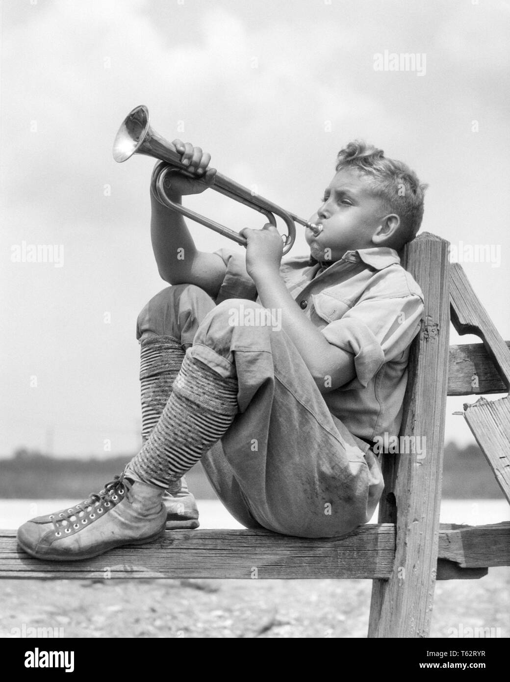 1930er Jahre jugendliche JUNGE IN KNIE HOSEN SOCKEN UND TURNSCHUHE SITZEN  AUF SCHIENE ZAUN SPIELEN BLASEN BUGLE-b 7941HAR 001 HARS MESSING AUSDRÜCKE  B&W ERFOLG SCHIENEN UND WANGEN AUFREGUNG LAUT PRALL AUF JUGENDLICHE