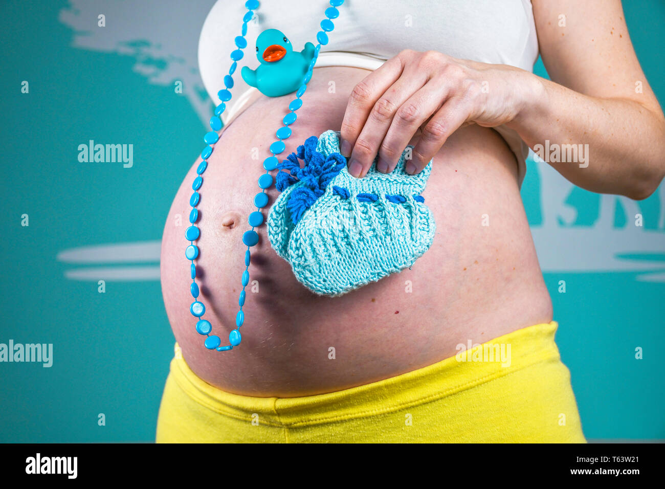 Junge schwangere Frau, ihren Bauch zu Hause zeigen schön abgerundet, indem die Hand blau Halskette, einem Kunststoff ente Spielzeug und gestrickte kleine Baby Schuhe Stockfoto