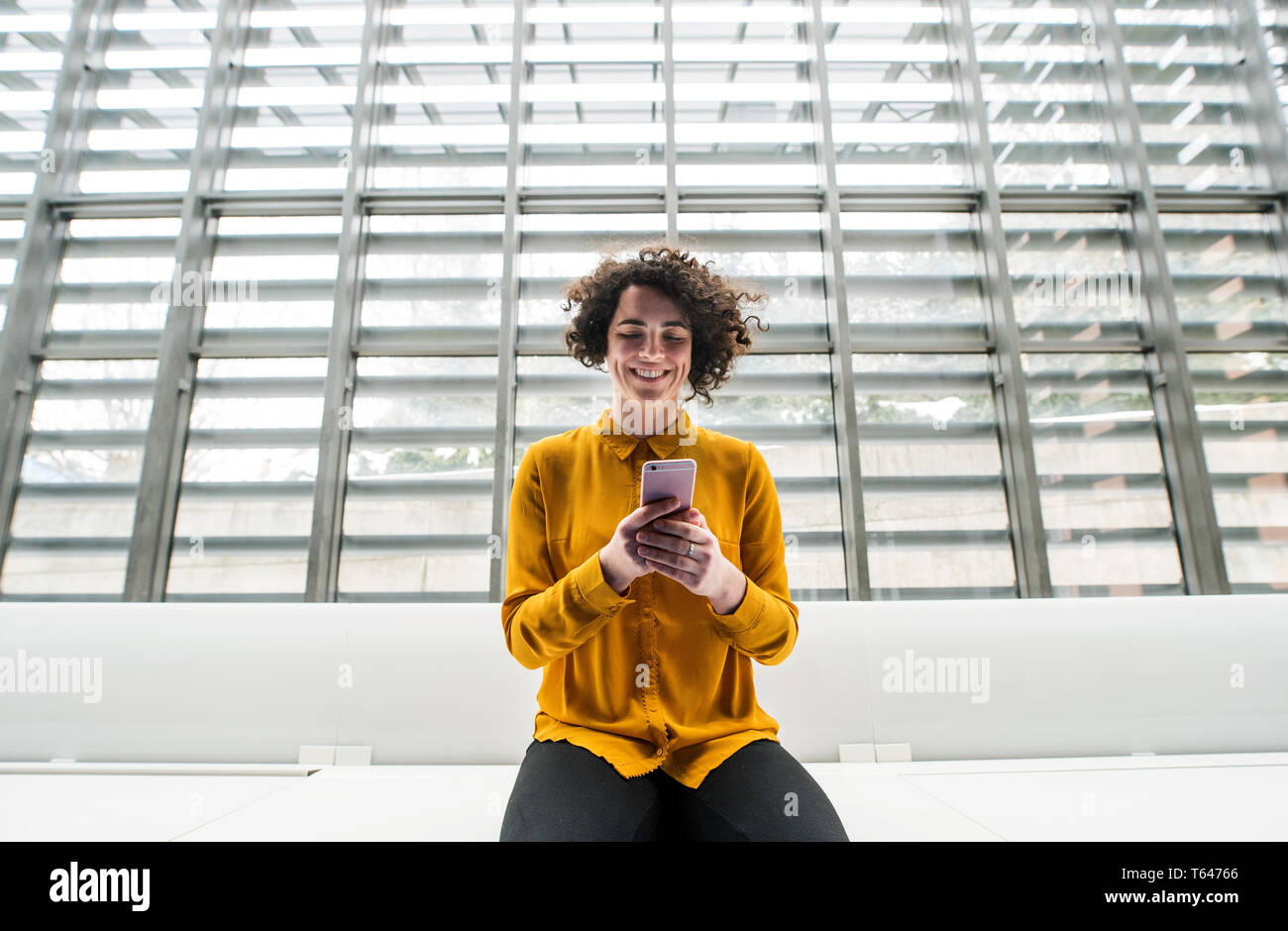 Das Porträt einer jungen Studentin oder Geschäftsfrau, sitzen am Schreibtisch im Zimmer in einer Bibliothek oder einem Büro, mit Smartphone. Stockfoto