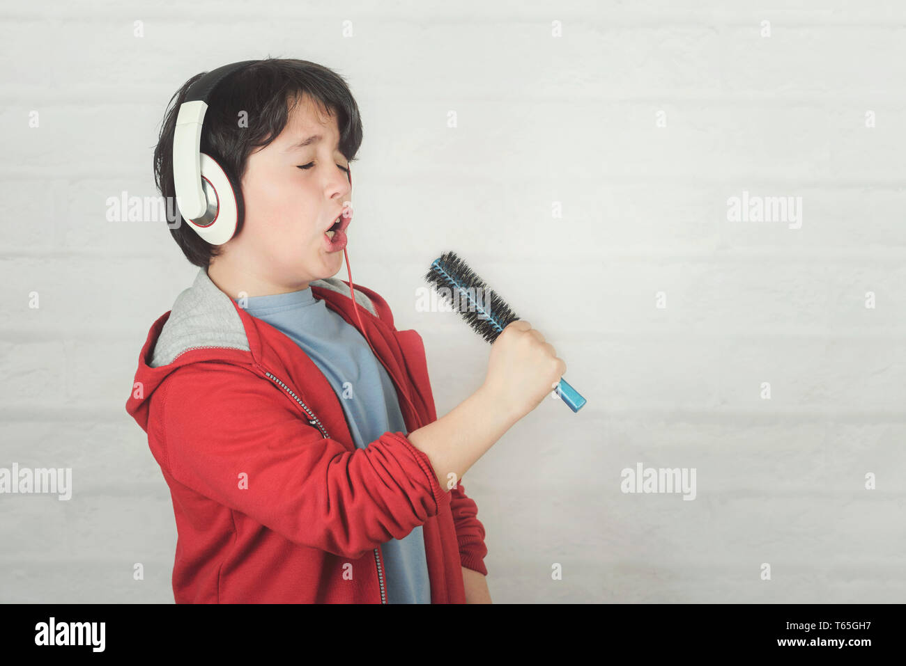 Lustig Kind in Kopfhörern singen mit Haar Bürste gegen Ziegel Hintergrund Stockfoto