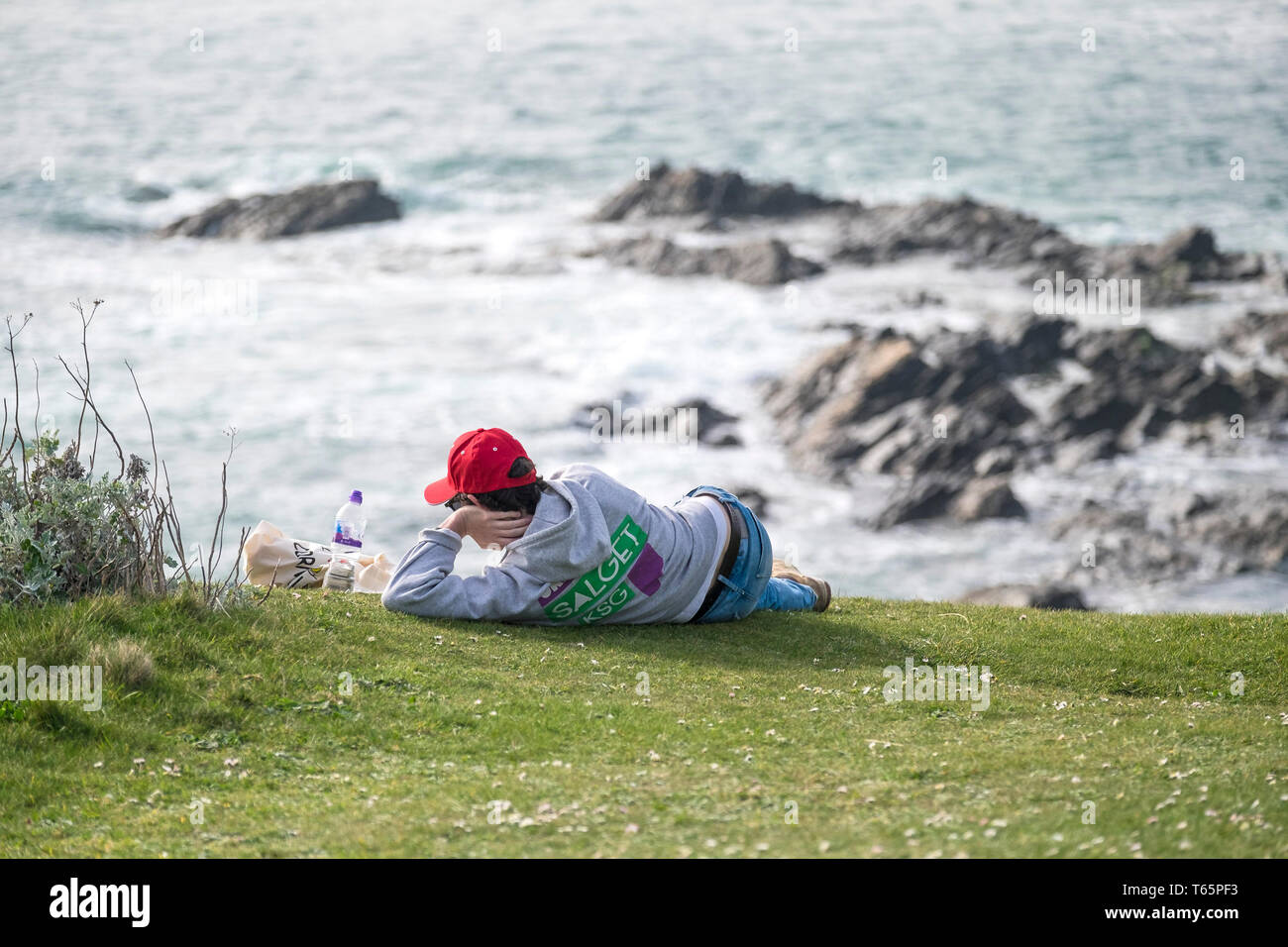 Ein Mann mit einem roten Baseball Cap und entspannend an der Küste mit Blick auf das Meer auf der Landspitze in Newquay in Cornwall. Stockfoto
