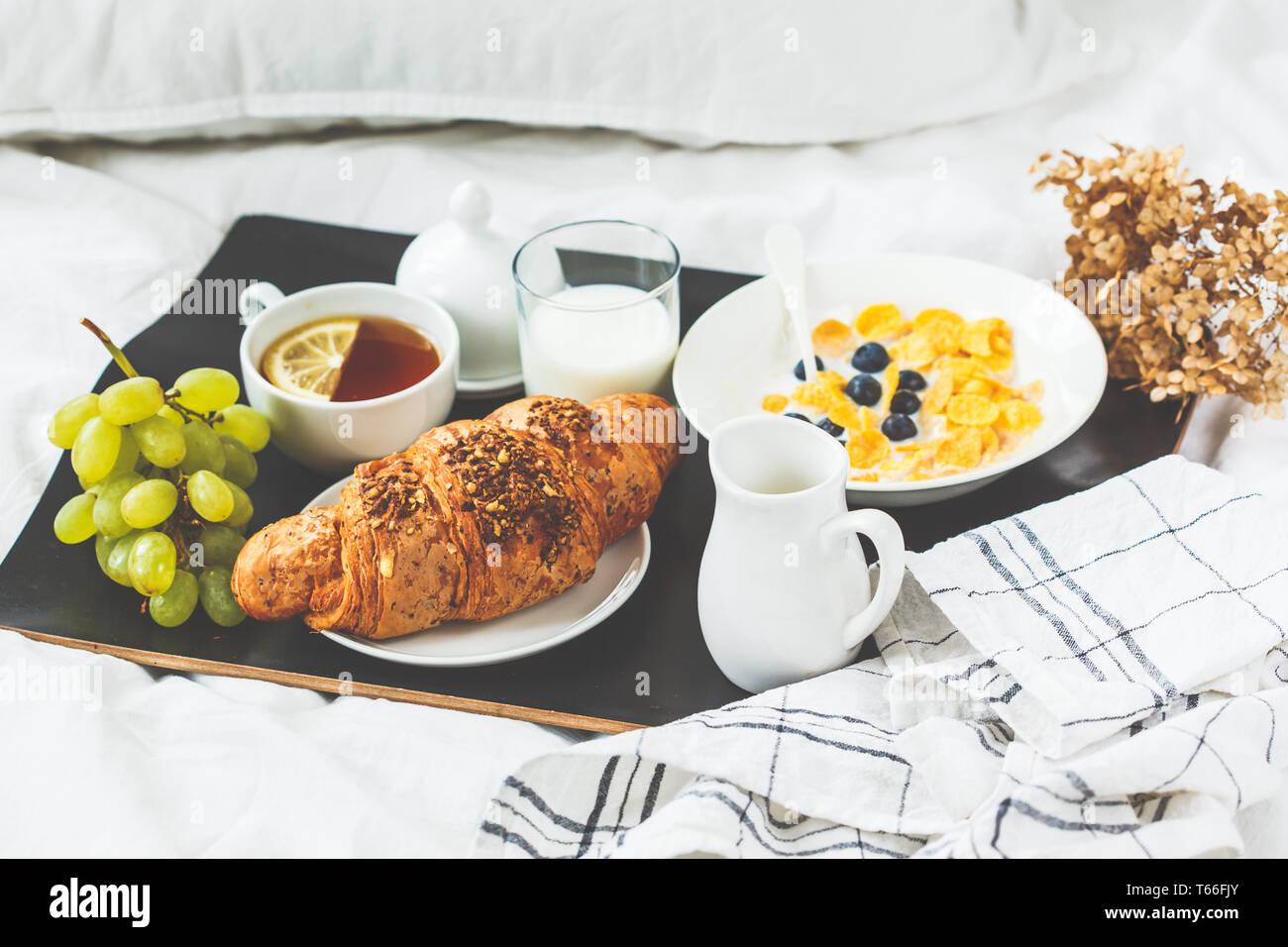 Kontinentales Frühstück im Bett. Croissant, Cornflakes, Tee, Milch und Obst auf einem schwarzen Fach. Stockfoto