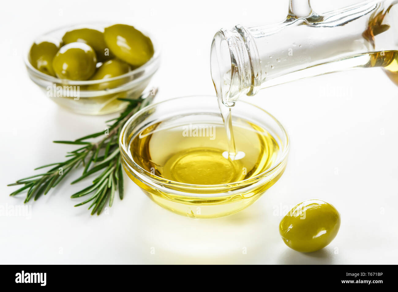 Olivenöl aus Glas gießen, frischen Oliven in Schalen- und Rosmarinzweige auf weißem Hintergrund Stockfoto