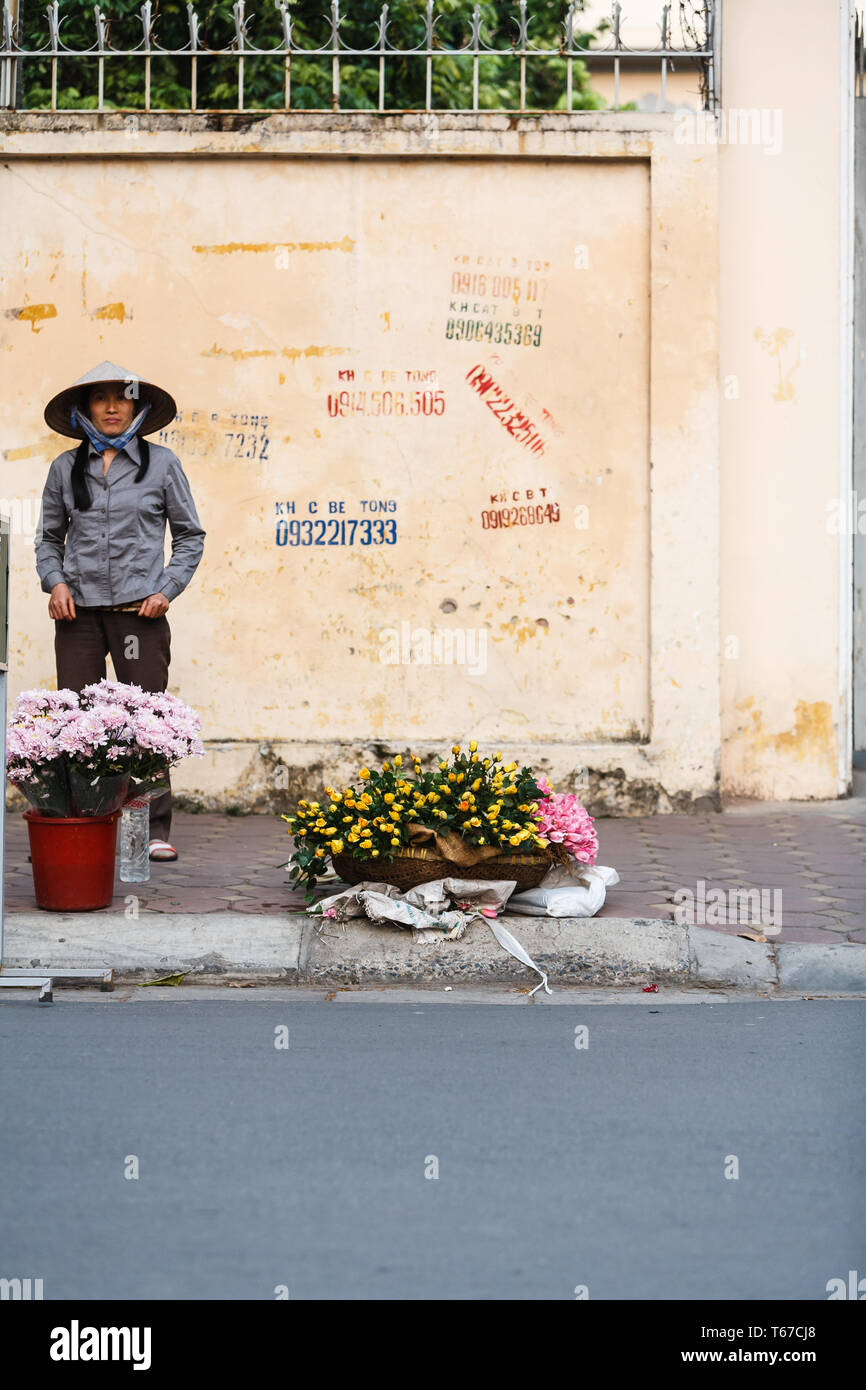 Hanoi, Vietnam, Frau, die Blumen auf Stadt Straße Ecke mit Wand hinter ihr mit Zahlen graffiti auf es Stockfoto