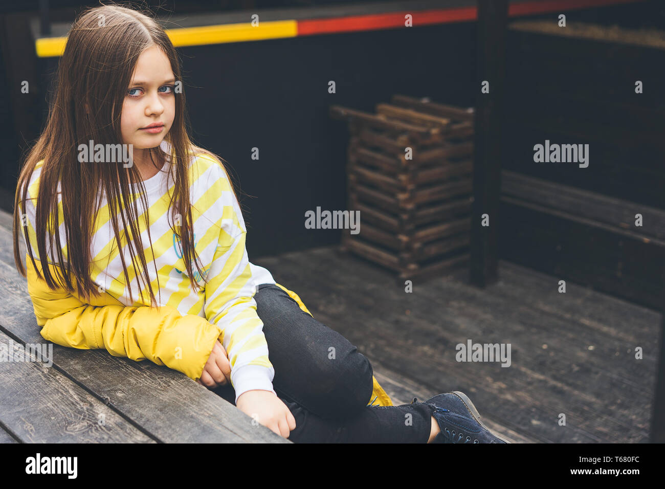 Jugendmädchen in gelb Pullover und schwarze Jeans sitzt auf einem Holz- Struktur in einem stadtbild Stockfoto