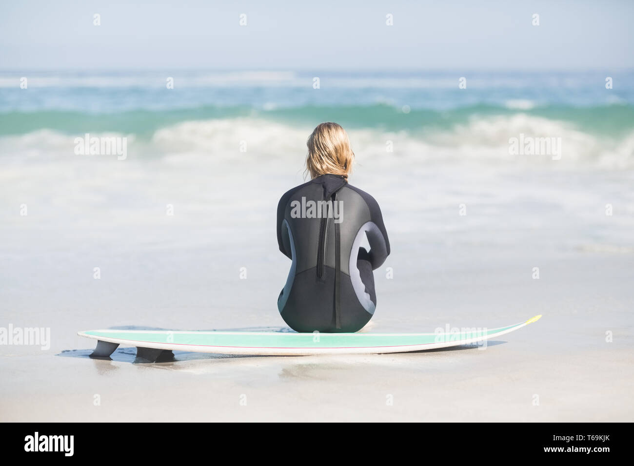 Rückansicht der Frau im Neoprenanzug mit Surfbrett am Strand sitzen Stockfoto
