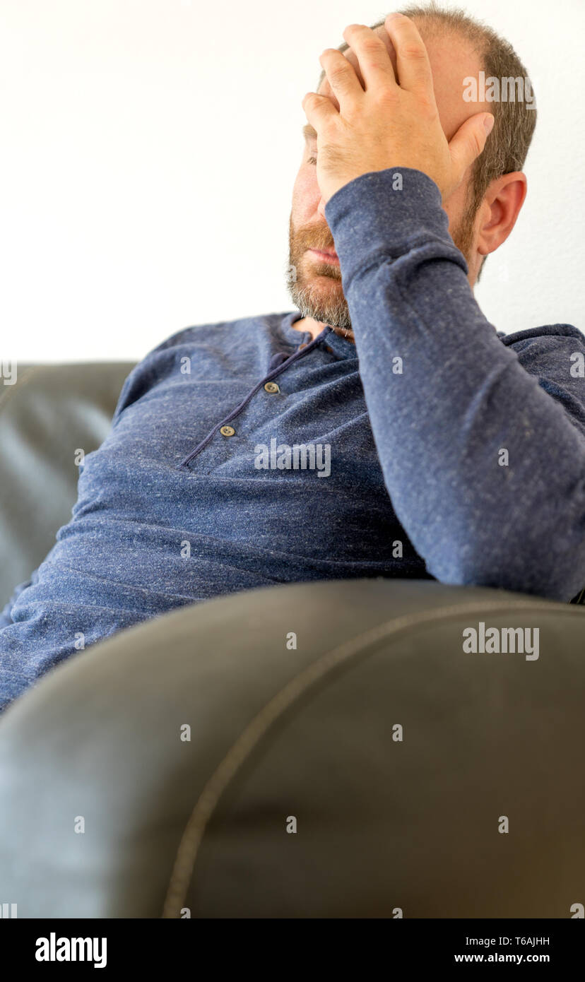 Erschöpft Mann mittleren Alters mit einem Bart sitzt auf dem Sofa und ruht sein müdes Haupt in seiner Hand. Stockfoto