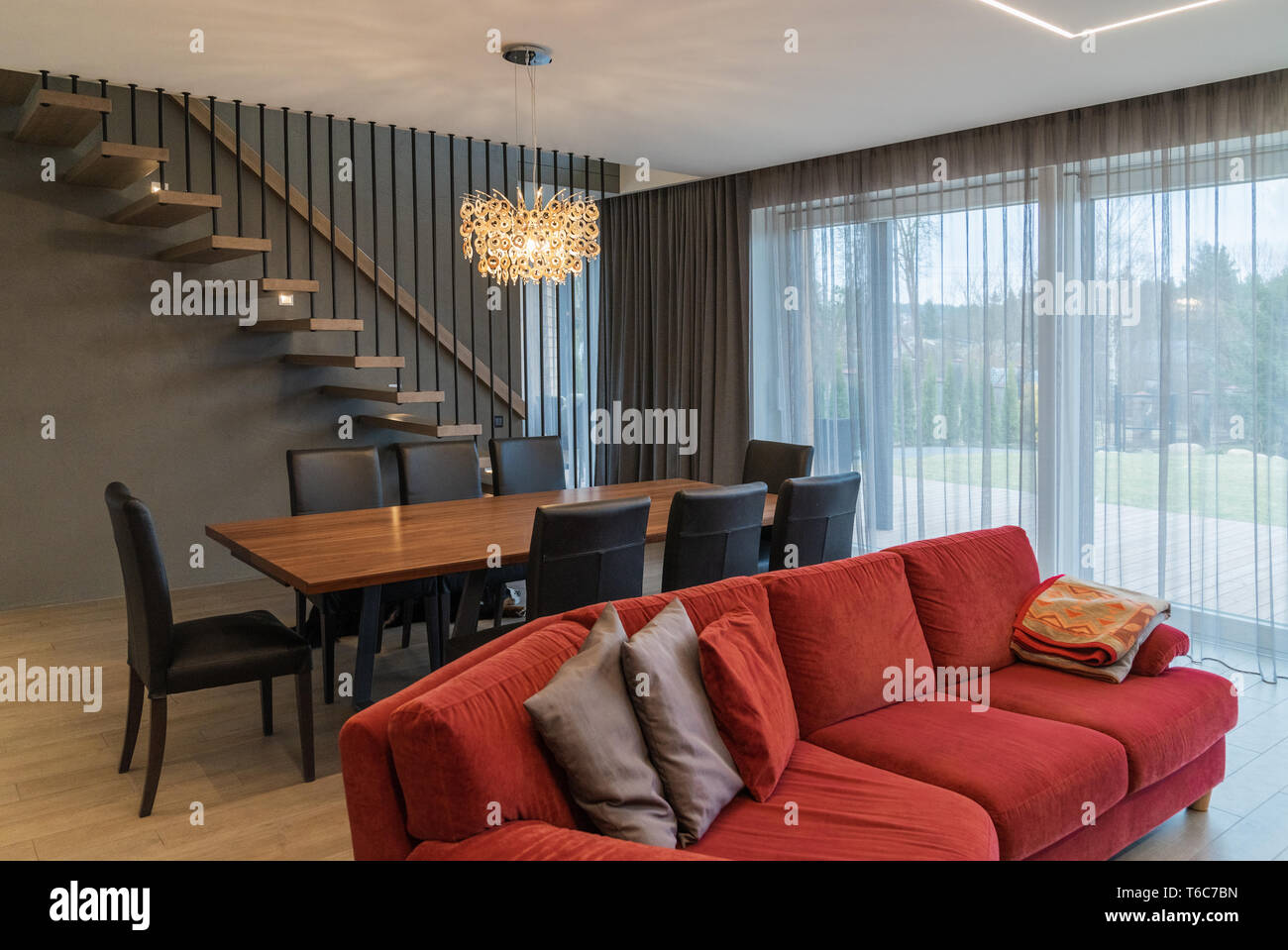 Essbereich und Wohnzimmer im Inneren des modernen Haus. Gemütliche Einrichtung mit roten Sofa, Esstisch, Stühle, Fenster, Lampe, und Treppenhaus. Stockfoto