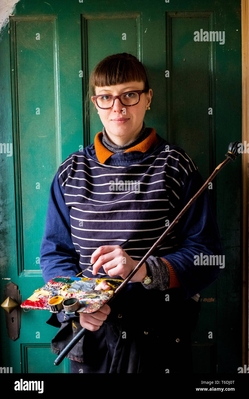 Frau artist Brille holding Pinsel, Palette und Mahlstick, an der Kamera schaut. Stockfoto