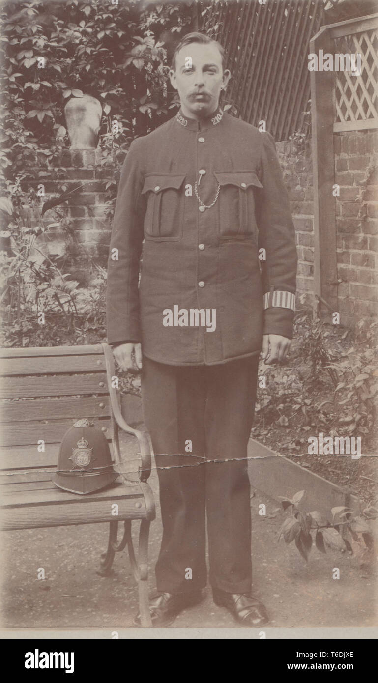 Jahrgang fotografische Postkarte zeigt ein Britischer Polizist stand in einem Garten mit seinen Helm auf einer Bank. Kragen Nr. M210 Stockfoto