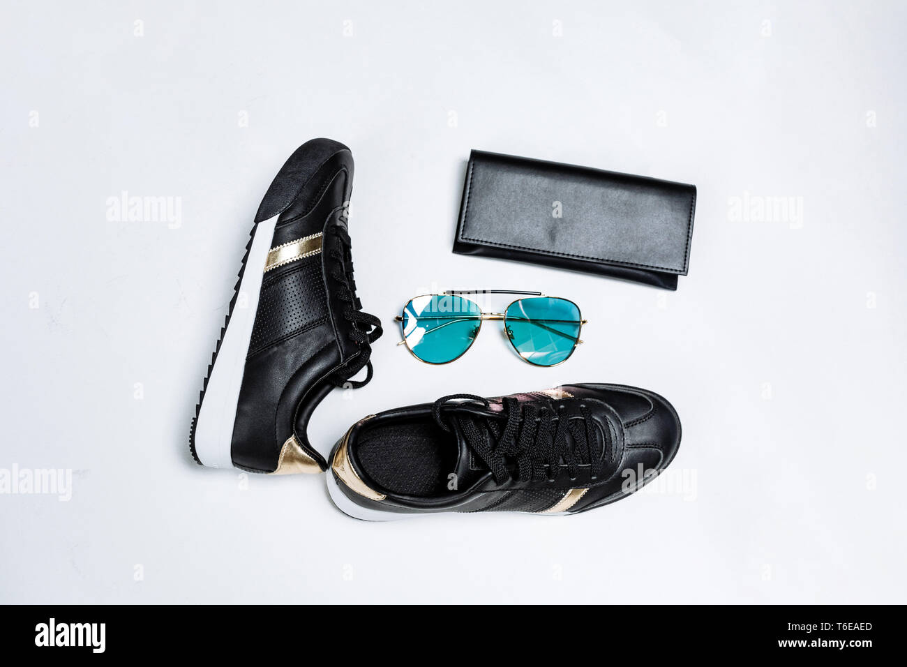 Modische schwarze Sneakers auf eine weiße Sohle mit schwarzen Socken und goldenen Akzenten, Sonnenbrille mit blauen Gläsern und eine schwarze Geldbörse auf weißem Hintergrund Stockfoto
