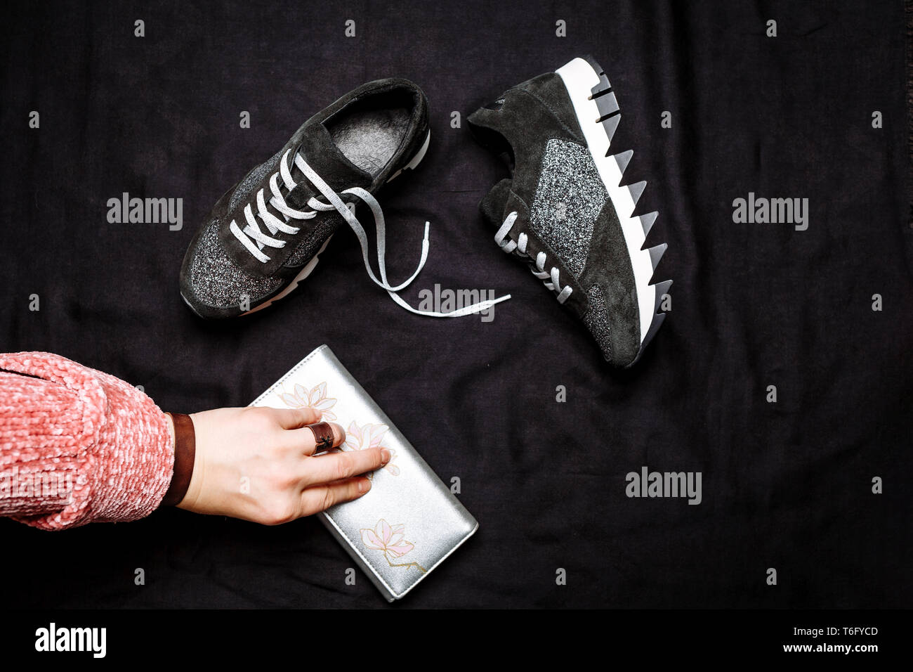 Schwarz Leder Sneakers mit silbernen Pailletten und eine weibliche Hand mit einem silbernen Geldbeutel auf einen schwarzen Woven Hintergrund altmodisch eingerichtet Stockfoto