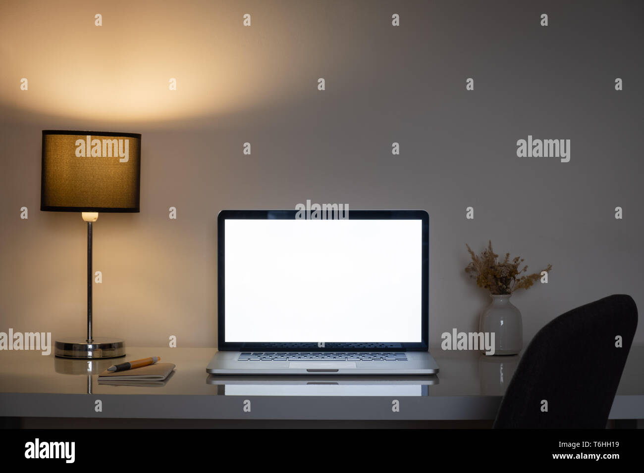 Gemütlich zu Hause Büro Schreibtisch in der Nacht beleuchtet durch eine Lampe und weißer Laptop Bildschirm. Saubere, moderne Arbeitsplätze für Telearbeit oder freiberufliche Tätigkeit, Low Key Stimmung Stockfoto