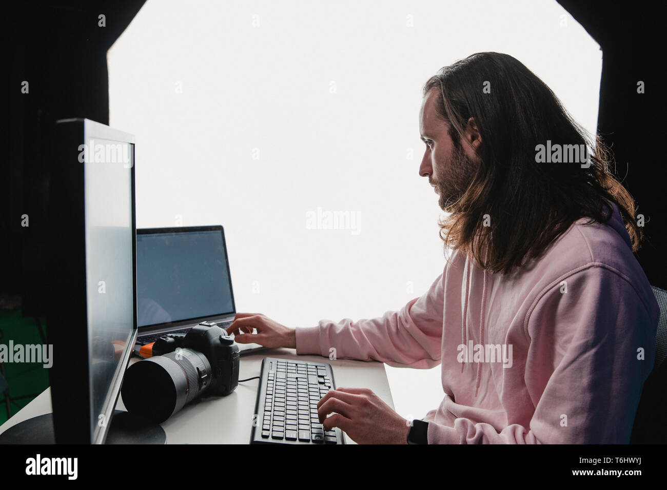 Eine Nahaufnahme Seite geschossen von einem kaukasischen Mann an seinem Schreibtisch in einem Film Studio arbeitet, einen Computer, Laptop und Kamera auf dem Schreibtisch gesehen werden kann, die junge ma Stockfoto