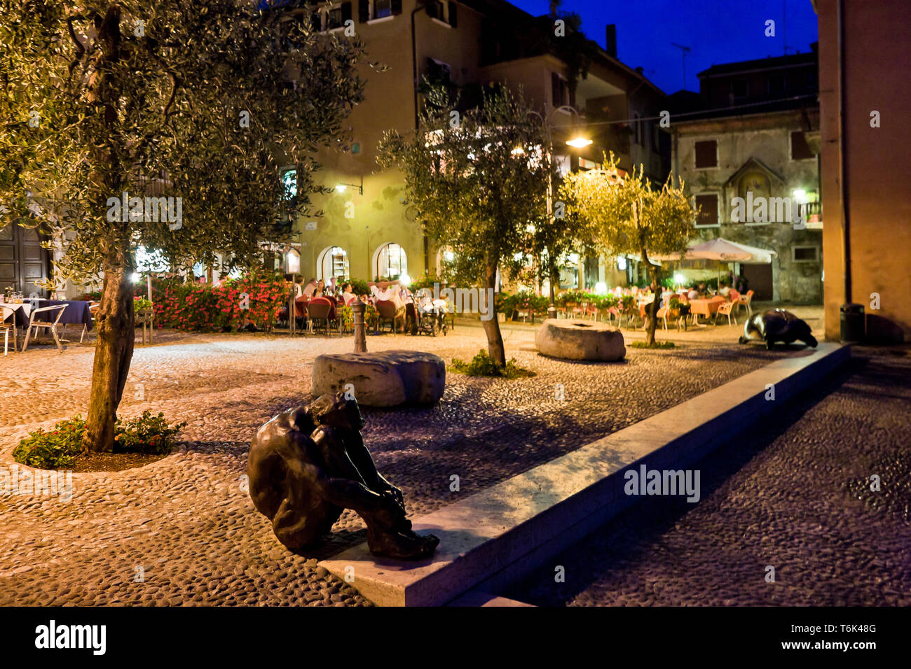 Malcesine: veduta serale di Piazza Porto Vecchio con Ulivi e l'Opera di Victor Ochoa intitolata 'El Zulo'. [ENG] Malcesine: Nacht Blick auf Porto Vecch Stockfoto